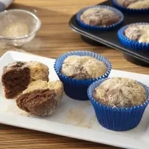 Foto da receita de muffin de cacau com farinha láctea. Observa-se dois muffins em forminhas azuis com farinha láctea polvilhada. Ao lado esquerdo, um muffin sem forminha cortado ao meio.