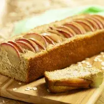 Fotografia em tons de marrom de uma bancada de madeira, sobre ela uma tábua de madeira com o bolo de maça. Ao fundo uma maçã e um paninho verde.