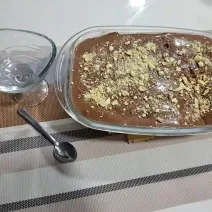 Foto da receita de Pavê de Cupuaçu com Ganache de Chocolate. Observa-se uma travessa retangular com o doce decorado de biscoito picado por cima e ao lado esquerdo tem uma taça individual.