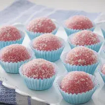 Fotografia em tons de azul e rosa de um paninho azul, sobre ele um prato redondo com decoração de florezinhas, sobre ele 12 brigadeiros rosa, com açúcar polvilhado, em forminhas azuis claras para doce.