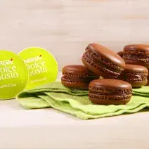 Fotografia em tons de verde em uma bancada de madeira clara, um paninho verde com vários macarons de chocolate e café em cima dele. Ao lado, cápsulas de Cappuccino Dolce Gusto.