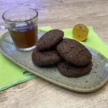 Fotografia em tons de verde em uma bancada de madeira, um recipiente oval marrom ao centro com 4 cookies de chocolate com amêndoas e ao lado uma xícara de vidro com o chá mate com acerola. Ao lado, uma cápsula de Dolce Gusto.
