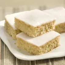 Fotografia em tons de bege em uma mesa de madeira com uma toalha bege listrada, um prato branco quadrado com pedaços de bolo de camomila com cobertura de glacê branco.