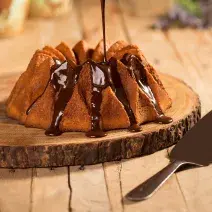 Foto aproximada de um bolo de cenoura sendo decorado com uma calda de chocolate, sobre uma tábua de madeira com uma espátula de corte ao lado