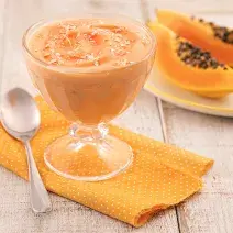Foto em tons de laranja da receita de creme de papaia servida em uma taça de vidro sobre um pano laranja em uma mesa de madeira. Ao lado há um colher prateada e ao fundo há um prato com duas fatias médias de mamão