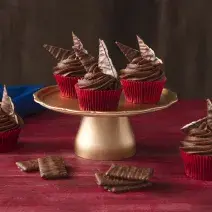 Foto de uma bancada vermelha. Ao centro há um suporte para bolos dourado alto com 3 cupcakes em formas vermelhas e decorados com Nestlé ChocoBiscuits. Na bancada há mais 2 cupcakes, alguns biscoitos e um tecido azul escuro ao fundo.