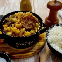 Foto da receita de Grão de Bico com Carnes. Observa-se uma mini panela com grão de bic e linguiça com carne e, ao lado direito, uma mini panela com arroz.