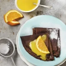 Fotografia de dois crepes de chocolate dobrados recheados com brigadeiro. Por cima, uma calda de laranja por cima, açúcar de confeiteiro polvilhado e meia rodela de laranja. Os crepes estão em um prato azul claro, e ao lado, mais calda de laranja.
