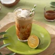 Foto da receita de iced coffee coco com caramelo servida em um copo de vidro alto com um canudo listrado verde em cima de um prato verde sobre uma tábua de madeira