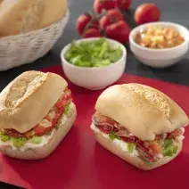 Foto de um sanduíche pequeno, recheado com uma creme branco, alface e tomate, sobre uma tábua vermelha em uma mesa com potinhos com ingredientes e um cesto de pães franceses ao fundo