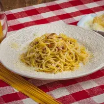 Foto de uma mesa com uma toalha xadrez vermelha e branca. Sobre ela há um prato branco com a receita de Spaghetti à Carbonara, alguns macarrões crus espalhados e num pote e, do lado direito, um pedaço de queijo com queijo ralado.