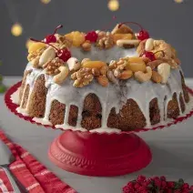 Fotografia em tons coloridos em uma bancada de madeira de cor cinza. Ao centro, uma boleira vermelha contendo o bolo e ao lado, um pano vermelho listrado e ao fundo, enfeites de natal.