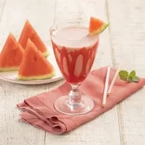 Foto da receita da bebida refrescante de melancia e morango servida em uma taça de vidro com um pedaço de melancia para decorar, há um pano vermelho embaixo da taça e sobre a mesa de madeira clara. Ao fundo um pires branco com três pedaços de melancia