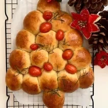 Foto da receita de Pão Caseiro Natalino. Observa-se um pão assado e dourado em formato de árvore, decorado com tomatinhos cereja