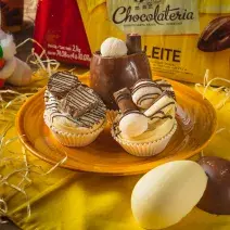 Foto aproximada de três miniovos de chocolate brancos, em um prato amarelo sobre uma bancada de madeira decorada com palha, um tecido amarelo, um pequeno coelho de pelúcia e uma barra de chocolate ao fundo