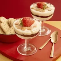 Foto da receita de Panacota de Frutas Vermelhas e Biscoito na taça. Observa-se duas taças com camadas e decoradas com lâminas de amêndoas e um morango por cima. Ao lado esquerdo, um potinho com biscoitos inteiros.
