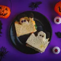 Imagem de um ângulo de cima. Numa mesa em tom roxo escuro há um prato preto com dois sanduíches de pão de forma cortados em formato de fantasmas, com olhos pretos. Ao redor há abóboras, morcegos e aranhas de mentira.