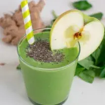 Fotografia em tons de verde em uma mesa branca com um copo de vidro alto e o smoothie de maçã verde com banana e chia dentro dele, com uma fatia de maçã enfeitando o copo. Ao fundo, folhas de espinafre e gengibre.
