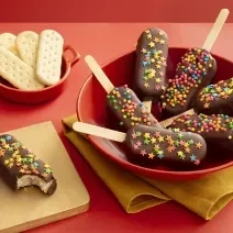Foto da receita de Pirulito de Biscoito. Observa-se 7 pirulitos decorados com chocolate e confeitos em um prato fundo vermelho sobre um guardanapo de pano amarelo