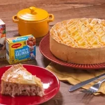 Foto da receita da Melhor Torta de Frango. Observa-se uma torta de frango redonda e grande e, à frente, em um prato de sobremesa, uma fatia dela. Embalagens de MAGGI decoram a foto.