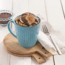Foto da receita de bolo de caneca com cacau e banana em uma caneca azul, sobre tábuas de madeira, com um guardanapo de pano branco e um garfo sobre ele.