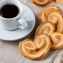 Foto da receita de Palmier ou Orelha de Macaco. Observa-se o biscoito em disposto em cima de um pano de prato e, ao lado, uma xícara com café sobre um pires.