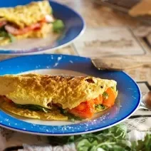 Fotografia em tons de azul e laranja em uma bancada de madeira escura, um prato redondo azul com uma omelete em cima dele recheada de tomate e rúcula. Ao lado colher e uns guardanapos marrons desenhados.