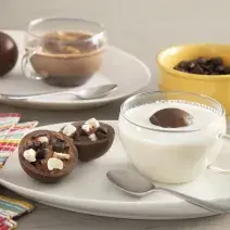 Fotografia em tons de branco e amarelo de uma bancada cinza, ao centro um prato com uma xícara com leite e o chocobomb e ao lado um chocobomb aberto, mostrando a camada de chocolate e o recheio.