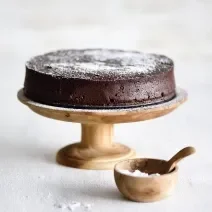 Foto da receita de Bolo Chocolatudo Sem Farinha. Observa-se uma boleira de madeira em um fundo branco com o bolo em cima, polvilhado de açúcar.