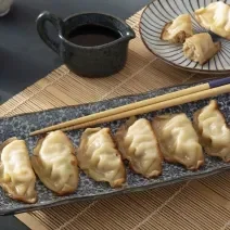 Foto da receita de Guioza. Observa-se 6 guiozas sobre uma peça de cerâmica asiática com um hashi apoiado. Atrás, um pratinho com duas guiozas e um potinho de shoyu ao lado.