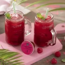 Foto em tons de cor de rosa da receita de pink lemonade com frutas vermelhas e hortelã servida em canecas altas de vidro em cima de uma tábua rosa clarinha com um pote de framboesas ao lado