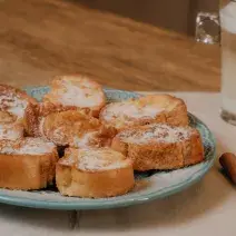 Fotografia em tons de marrom de uma bancada de madeira com um paninho branco sobre ele um prato azul claro e rabanas com açúcar e canela. Ao fundo paus de canela com um copo de leite.