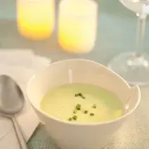 Fotografia em tons de verde em uma mesa de madeira com uma toalha verde, ao centro um potinho branco redondo fundo com o creme de abobrinha com cebolinha decorando. Ao fundo, duas velas acesas e uma taça de vidro com água.