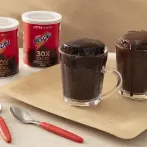 Foto da receita de Bolo de chocolate na caneca servida em duas canecas de vidro sobre uma tábua de madeira com duas latas de nescau extra cacau e duas colheres de decoração