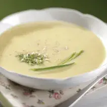 Fotografia em tons de verde em uma bancada com uma bandeja branca, um pano branco com detalhes verde e em flores, um prato redondo branco fundo em cima dela e o creme de mandioquinha.