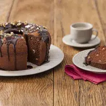 Fotografia em tons de marrom em uma mesa de madeira com um prato branco grande e o bolo de chocolate e nozes e ao lado um prato branco pequeno com uma fatia do bolo. Ao fundo, um pires e uma xícara branca.