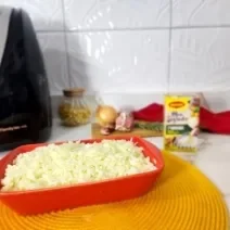 Foto da receita de arroz na airfryer servida em uma travessa vermelha com uma airfryer ao fundo junto com uma embalagem de maggi meu segredo, cebolas e um pano vermelho