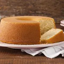 Fotografia em tons de marrom, laranja e branco de uma bancada marrom vista de frente, um prato branco contém um bolo com uma fatia cortada ao lado e abaixo e um pano branco. Ao fundo dois pratos brancos com uma fatia por cima e uma jarra branca.