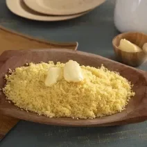 Foto da receita de Cuscuz de Milho. Observa-se uma tigela de madeira com o cuscuz soltinho e com manteiga em cima derretendo.