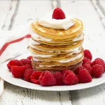 Fotografia em tons de branco em uma bancada de madeira de cor branca. Ao centro, um prato branco contendo as pancakes com frutas espalhadas ao redor. Ao lado, um pano branco espalhado.