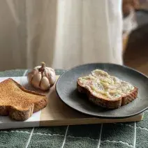Foto de um prato escuro redondo com uma fatia de pão com a manteiga de alho por cima. Ao lado esquerdo dele há outra fatia de pão torrada e um dente de alho inteiro ao fundo.