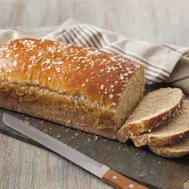 Em uma mesa de madeira contém um pão com a metade fatiado. Ao lado um pano nas cores cinza e marrom e do outro lado uma faca.