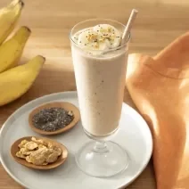 Foto da receita de Smoothie de Banana com Nesfit Cereal. Observa-se um copo alto com o smoothie dentro e finalizado com NESFIT Cereal, banana e chia. Ao lado esquerdo, potinhos com NESFIT cereal, chia e um cacho de bananas decoram a foto.