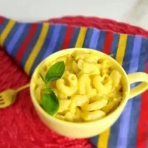Foto da receita do mac and cheese na caneca servida em uma caneca amarela sobre um pano listrado colorido e ao lado um garfo dourado