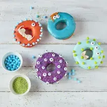 Imagem vista de cima de alguns Donuts Monstro em tons coloridos, decorados com tema de dia das bruxas, tudo em uma bancada de madeira em tom claro