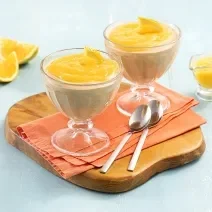 Foto da receita de Mousse de Cappuccino com Calda de Laranja. Oberva-se duas taças transparentes com a mousse e a calda laranja por cima em cima de um guardanapo laranja e uma tábua redonda de madeira. Como decoração, pedaços de laranja em um fundo azul.