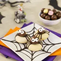 Imagem aproximada de um prato quadrado branco com três biscoitos em formatos de aranhas. A bancada está decora com papéis em tons roxo e laranja e alguns morcegos de mentira.