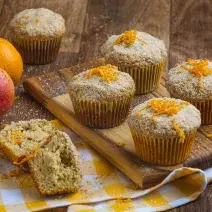 Imagem aproximada de muffins em tom claro, sobre uma tábua de madeira em um pano quadriculado em tom amarelo e branco. Ao fundo há uma maçã e uma laranja e alguns farelos de Farinha Láctea.
