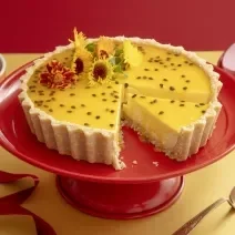 Foto da receita de Cheesecake de Maracujá e Chocolate Branco. Observa-se uma torta com calda de maracujá e flores comestíveis com um pedaço cortado, em cima de uma boleira vermelha.