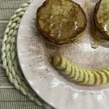 Foto da receita de Panquequinha de Aveia. Observa-se um prato com panquequinhas com mel e canela e do lado de baixo do prato, uma banana em rodelas para formar um sorriso.
